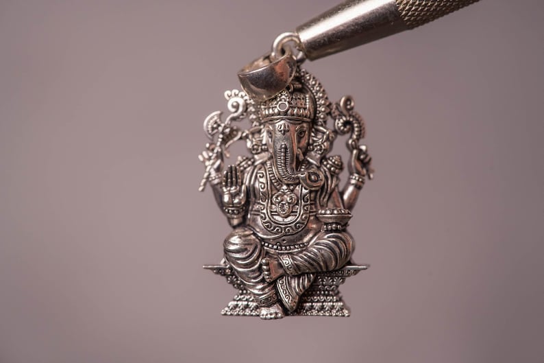 May include: A detailed silver pendant depicting the Hindu deity Ganesha, seated with multiple arms and an elephant head. The pendant is intricately designed with ornate details and is suspended from a silver bail.