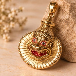 May include: A gold-toned metal pendant featuring a detailed relief of a deity with a fierce expression, sharp teeth, and red enamel accents around the eyes and mouth. The pendant has a circular, fluted border and a top bail for hanging.