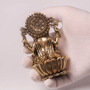 May include: A golden statue of a deity, seated on a lotus flower. The statue has intricate details, including a halo and ornate jewelry. The figure is held in a gloved hand against a white background.