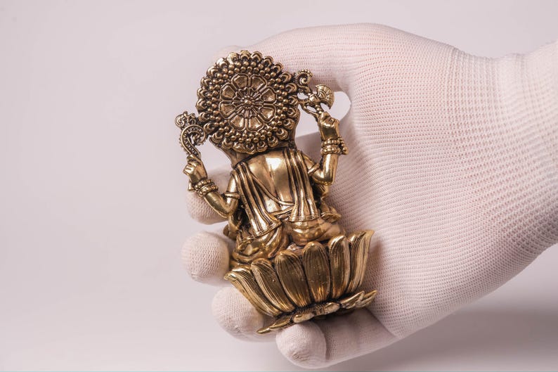 May include: A golden statue of a deity, seated on a lotus flower. The statue has intricate details, including a halo and ornate jewelry. The figure is held in a gloved hand against a white background.