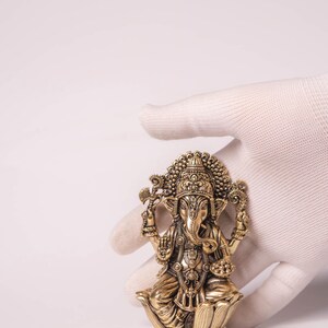 May include: A detailed brass statue of the Hindu deity Ganesha, seated on a lotus flower. The statue features intricate carvings, a large head, and multiple arms, held by a gloved hand against a white background. The statue is a golden color.