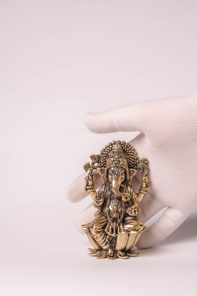 May include: A detailed brass statue of the Hindu deity Ganesha, seated on a lotus flower. The statue features intricate carvings, a large head, and multiple arms, held by a gloved hand against a white background. The statue is a golden color.