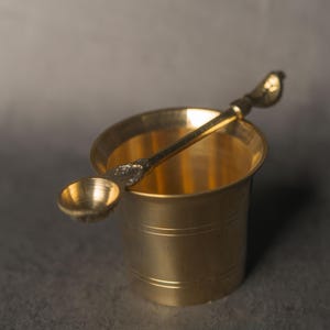 Könnte beinhalten: Eine kleine Messing-Tasse mit passendem Löffel. Die Tasse ist zylindrisch mit leicht ausgestelltem Rand. Der Löffel hat eine kleine, runde Schale und einen dekorativen Griff. Die Artikel sind aus glänzendem, goldenem Messing.