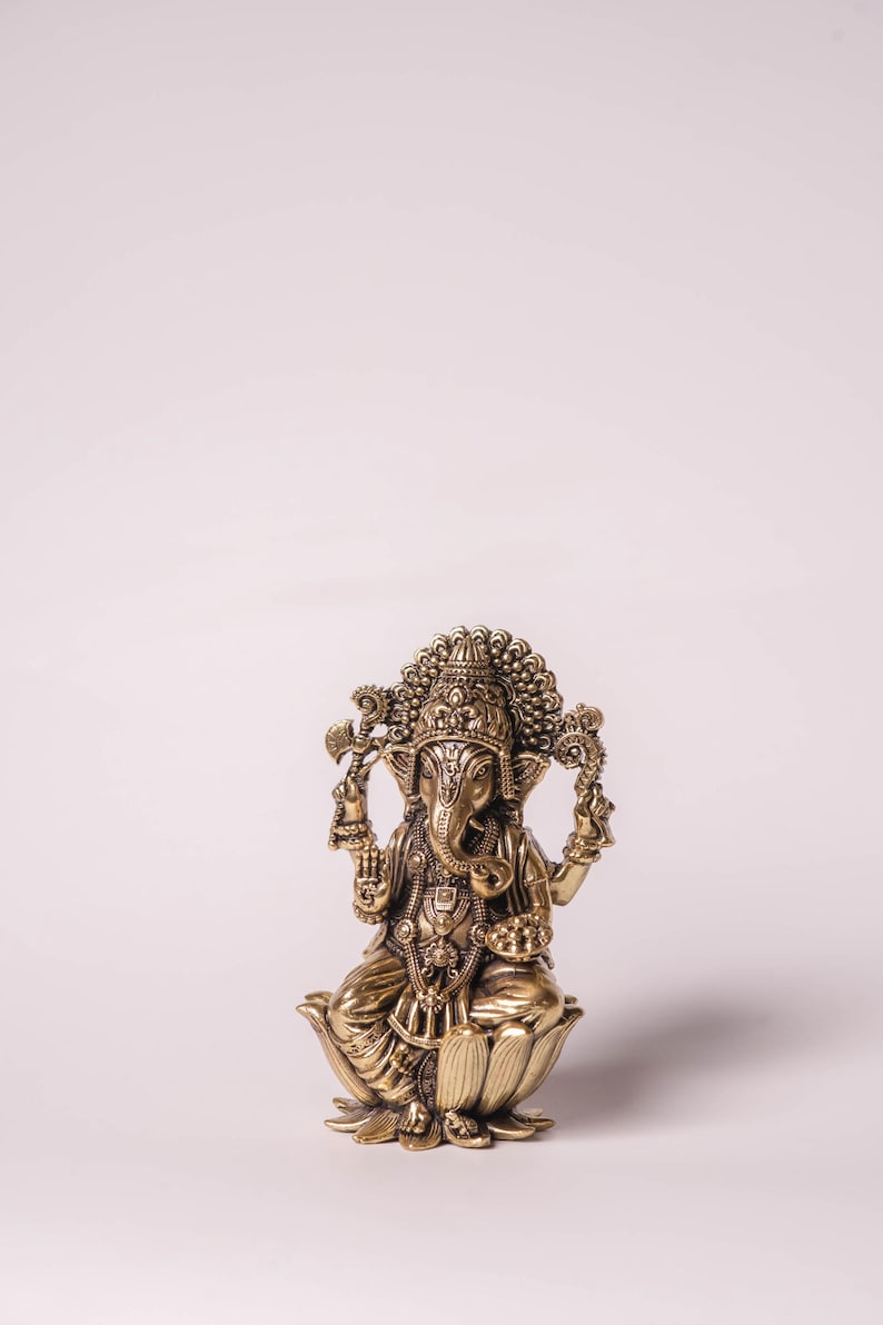 May include: A detailed brass statue of the Hindu deity Ganesha, seated on a lotus flower. The statue features intricate carvings, a large head, and multiple arms holding various objects. The figure is adorned with ornate jewelry and a crown.