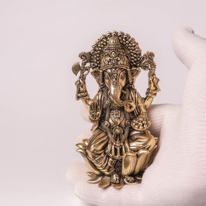 May include: A detailed brass statue of the Hindu deity Ganesha, seated on a lotus flower. The statue is held by a gloved hand, showcasing its intricate design and golden color. The figure has multiple arms and an elephant head.