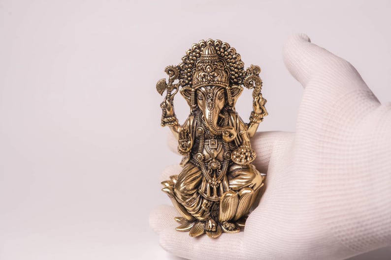 May include: A detailed brass statue of the Hindu deity Ganesha, seated on a lotus flower. The statue is held by a gloved hand, showcasing its intricate design and golden color. The figure has multiple arms and an elephant head.