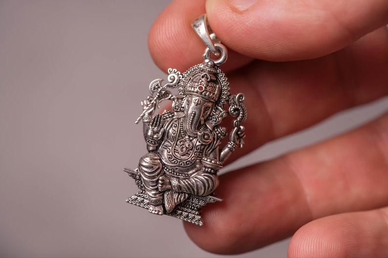 May include: A detailed silver pendant depicting the Hindu deity Ganesha. The pendant features intricate carvings and is held by a person's hand. The pendant has a loop for a chain and is a religious item.