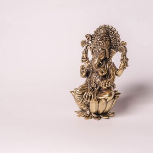 May include: A detailed bronze-colored statue of the Hindu deity Ganesha. The figure is seated on a lotus flower base, with multiple arms and an elephant head. Intricate details adorn the crown and clothing. The statue is a religious or decorative item.