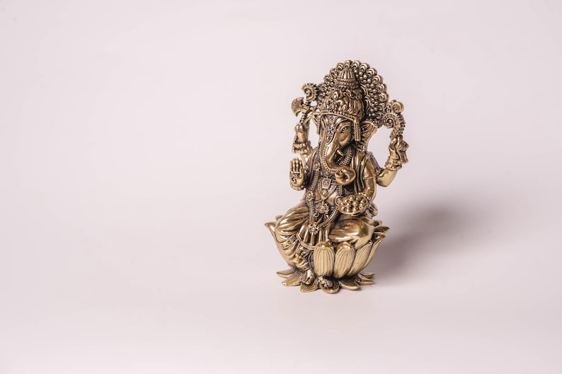 May include: A detailed bronze-colored statue of the Hindu deity Ganesha. The figure is seated on a lotus flower base, with multiple arms and an elephant head. Intricate details adorn the crown and clothing. The statue is a religious or decorative item.