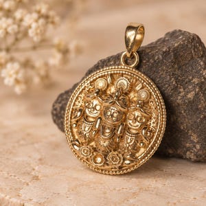 May include: A circular gold pendant featuring a detailed relief of three figures with large eyes and ornate headpieces. The pendant has a beaded border and a polished gold bail at the top, showcasing intricate metalwork and a warm, metallic finish.