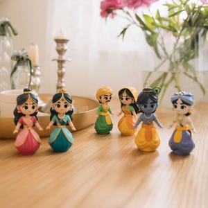 Op de afbeelding: Een verzameling kleurrijke beeldjes die traditionele Indiase personages uitbeelden. De beeldjes zijn gekleed in levendige kleding en versierd met sieraden. Ze zijn gerangschikt op een houten oppervlak, met een vaas met roze bloemen op de achtergrond.