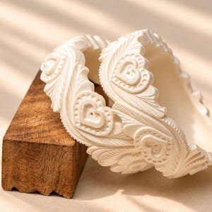 Puede incluir: Dos brazaletes de color blanco roto con un patrón en relieve de corazones y hojas estilizadas. Los brazaletes tienen un perfil grueso y redondeado con un acabado mate y se muestran apoyados contra un pequeño bloque rectangular de madera oscura.
