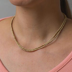 Puede incluir: Un delicado collar de varias capas con un diseño de cuentas doradas. El collar se lleva sobre una parte superior de color melocotón, creando un contraste sutil. Las cuentas parecen pequeñas y espaciadas uniformemente, formando un accesorio refinado.
