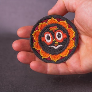 Op de afbeelding: Geborduurde patch met een zonachtig ontwerp. De patch heeft een zwart midden met een gezicht, omringd door oranje en gele bloemblaadjes en een bordeauxrode rand. De patch is rond en wordt in een hand gehouden.