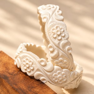 Puede incluir: Dos brazaletes de color blanco roto con un patrón floral y de remolinos en relieve. Las pulseras tienen un acabado mate suave y una forma circular consistente, mostrando tallas detalladas en relieve de pequeñas flores y líneas.