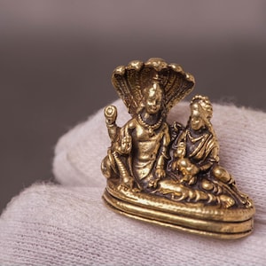 Könnte beinhalten: Kleine, goldfarbene Statue, die zwei Figuren darstellt, eine davon mit einer Kobrakappe, die zusammen sitzen. Die detaillierte Skulptur zeigt die Figuren in sitzender Position mit komplizierten Details an Kleidung und Schmuck.