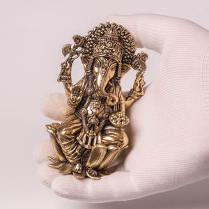 May include: A detailed brass figurine of the Hindu deity Ganesha, seated in a lotus position. The sculpture features intricate details, including a large elephant head, multiple arms, and ornate jewelry. The figurine is held in a gloved hand against a white background.