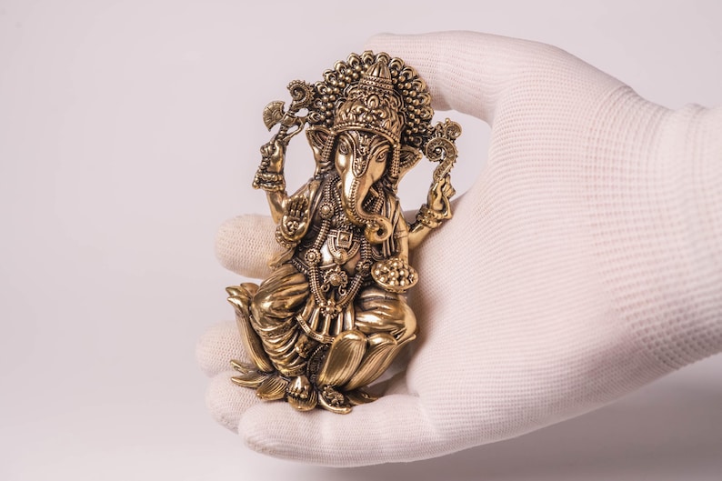 May include: A detailed brass figurine of the Hindu deity Ganesha, seated in a lotus position. The sculpture features intricate details, including a large elephant head, multiple arms, and ornate jewelry. The figurine is held in a gloved hand against a white background.