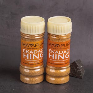 May include: Two spice jars with beige lids and orange labels. The labels read "MAYAPURE EKADASI HING" with text about being natural and preservative-free. A dark gray stone sits beside the jars.