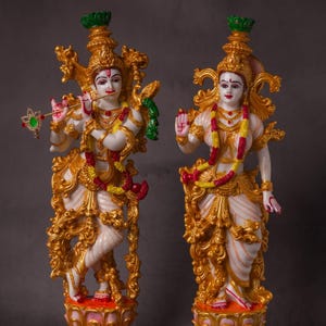 May include: Two ornate statues of Hindu deities. The figures are white with gold accents, elaborate headdresses, and jewelry. One holds a flute, the other a hand gesture. Both stand on detailed, floral-patterned bases. The statues are approximately 30 cm tall.
