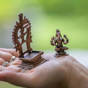 May include: Two small, bronze-coloured religious statues are displayed. One is a seated figure with multiple arms, and the other is a decorative wheel-like object. The statues are held in a hand against a blurred green background.