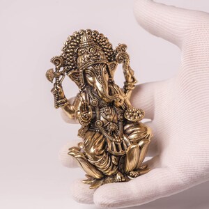 May include: A detailed golden statue of the Hindu deity Ganesha, held in a gloved hand. The statue features intricate carvings, a large head, and multiple arms, showcasing traditional Indian artistry. The figure is posed in a seated position.