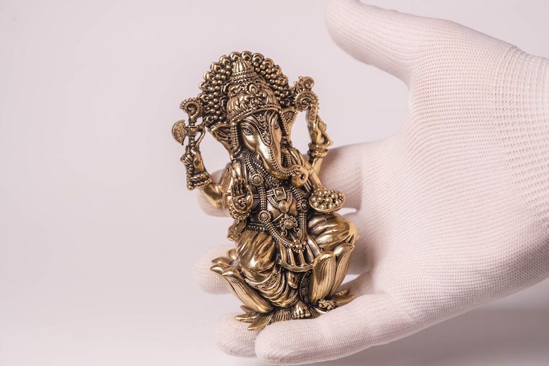 May include: A detailed golden statue of the Hindu deity Ganesha, held in a gloved hand. The statue features intricate carvings, a large head, and multiple arms, showcasing traditional Indian artistry. The figure is posed in a seated position.