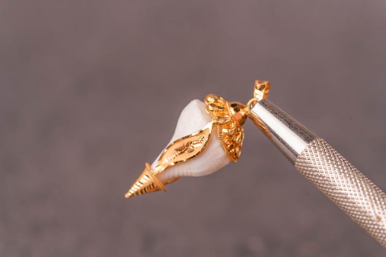 以下が含まれることがあります： 白い貝殻に金色の装飾が施されたゴールドのペンダント。ペンダントは銀色のツールで保持されており、ジュエリーの職人技を示しています。金のアクセントが貝殻の自然な形を際立たせています。
