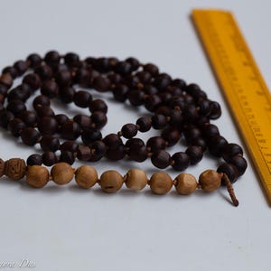 May include: Two strands of prayer beads with dark brown and light brown wooden beads. A yellow wooden ruler is positioned next to the beads. The beads are on a white surface.