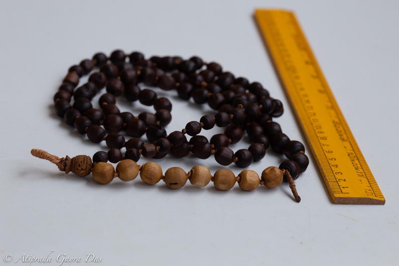 May include: Two strands of prayer beads with dark brown and light brown wooden beads. A yellow wooden ruler is positioned next to the beads. The beads are on a white surface.