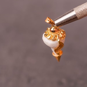 以下が含まれることがあります： 金色のペンダント。巻貝のデザインで、白い貝殻に金色のアクセントと装飾的な金のキャップが付いています。ペンダントは、灰色の背景に対して銀色のツールで保持されています。