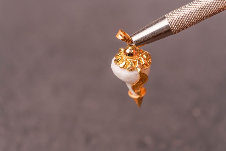以下が含まれることがあります： 金色のペンダント。巻貝のデザインで、白い貝殻に金色のアクセントと装飾的な金のキャップが付いています。ペンダントは、灰色の背景に対して銀色のツールで保持されています。