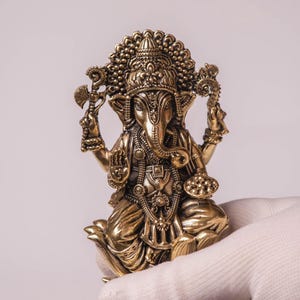May include: A detailed brass statue of the Hindu deity Ganesha, seated in a lotus position. The statue features intricate carvings, a large elephant head, and multiple arms holding various symbolic objects. The figure is held by a gloved hand against a plain background.