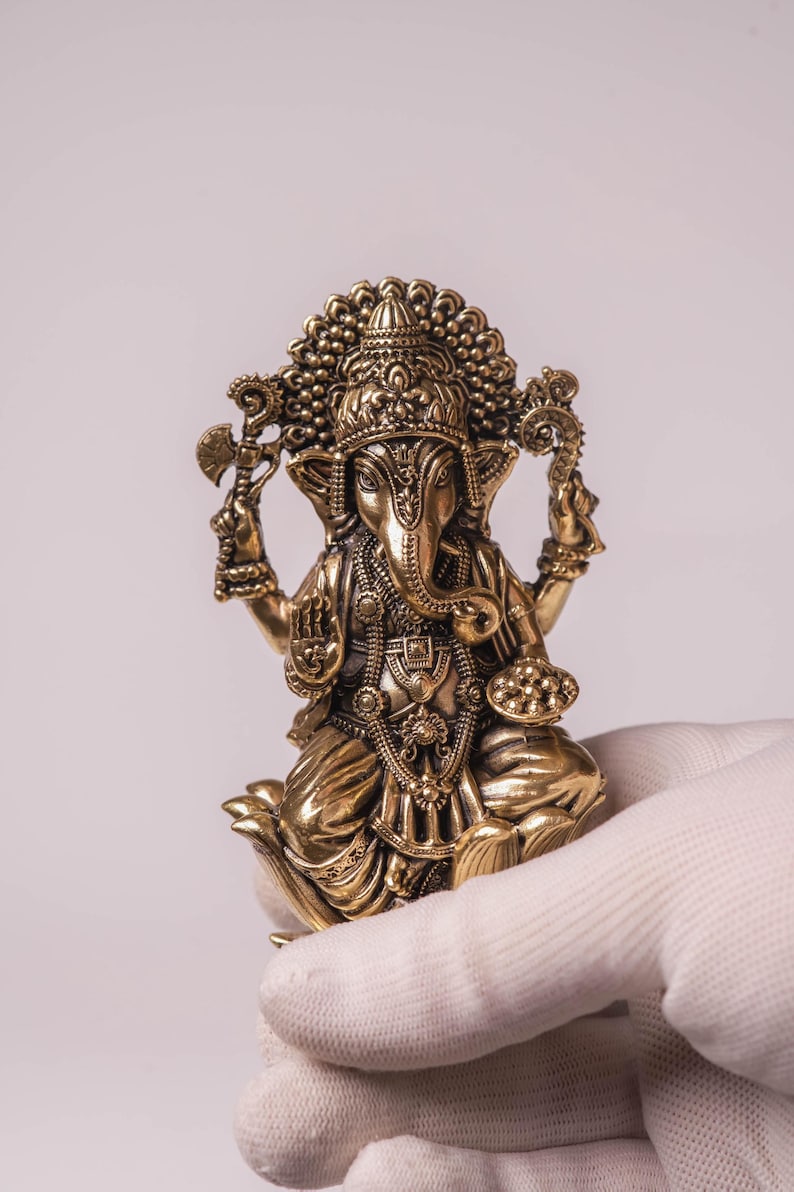 May include: A detailed brass statue of the Hindu deity Ganesha, seated in a lotus position. The statue features intricate carvings, a large elephant head, and multiple arms holding various symbolic objects. The figure is held by a gloved hand against a plain background.
