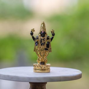 May include: A gold-colored statue of a deity with multiple arms, adorned with intricate jewelry and a tall headdress. The statue has a black body and is standing on a gold base. The background is a soft, blurred green.