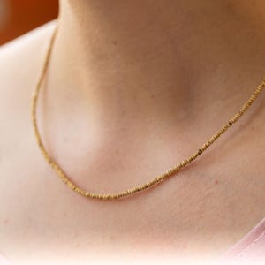 Puede incluir: Un delicado collar de cuentas doradas. El collar está compuesto por pequeñas cuentas espaciadas uniformemente, creando un diseño sutil y elegante. El collar se muestra sobre un fondo rosa suave.