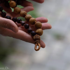 May include: A strand of prayer beads with alternating dark brown and light brown wooden beads. The beads are held in a person's hands, with a carved wooden pendant at the end. The background is blurred.