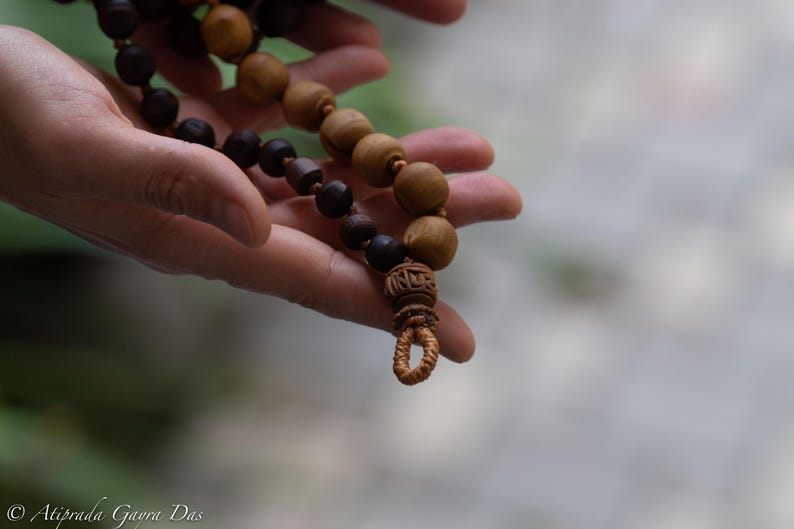 May include: A strand of prayer beads with alternating dark brown and light brown wooden beads. The beads are held in a person's hands, with a carved wooden pendant at the end. The background is blurred.
