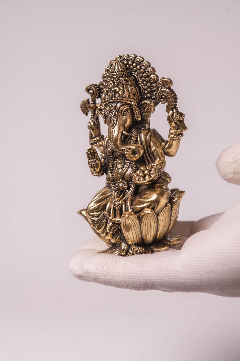 May include: A detailed brass statue of the Hindu deity Ganesha, seated on a lotus flower. The statue features intricate carvings, a large head, and multiple arms, with a detailed crown. The figure is held in a gloved hand against a white background.