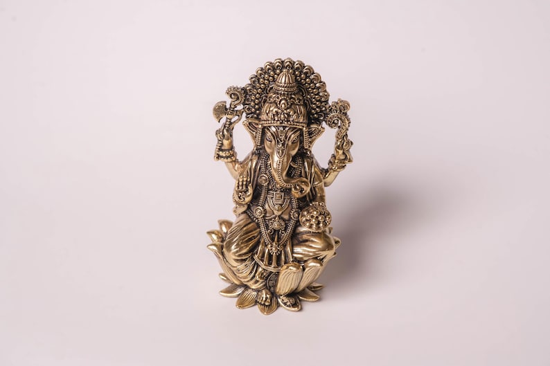 May include: A detailed brass statue of the Hindu deity Ganesha, seated on a lotus flower. The statue features intricate carvings, a large head with elephant features, and multiple arms holding various objects. The figure is adorned with ornate jewelry and a crown.