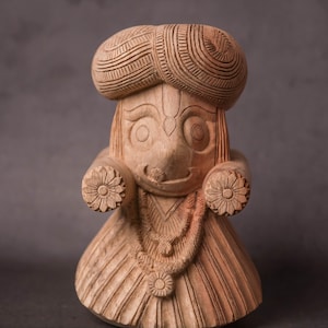 Puede incluir: Una figura de madera tallada de una figura con turbante, detalles florales y un collar. La figura tiene una expresión sonriente y está tallada en madera de color marrón claro. El fondo es gris liso.