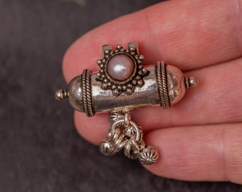 May include: A silver pendant with a central pearl, surrounded by a decorative sunburst design. The pendant has a cylindrical body with rope-like detailing and small dangling ornaments, showcasing intricate craftsmanship.