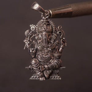 May include: A detailed silver pendant depicting the Hindu deity Ganesha, seated with multiple arms and an elephant head. The pendant is suspended from a silver bail, held by a tool against a dark background. The intricate design showcases traditional religious iconography.
