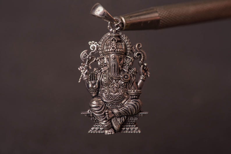 May include: A detailed silver pendant depicting the Hindu deity Ganesha, seated with multiple arms and an elephant head. The pendant is suspended from a silver bail, held by a tool against a dark background. The intricate design showcases traditional religious iconography.