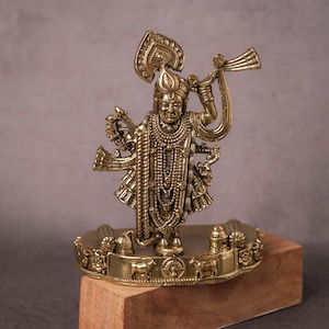 Op de afbeelding: Gedetailleerd messing beeld van een godheid, met meerdere armen en sierlijke sieraden. De figuur houdt een hoorn vast en is gemonteerd op een decoratieve basis met diermotieven. Het beeld staat op een houten blok tegen een neutrale achtergrond.