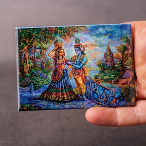 Op de afbeelding: Een rechthoekige kunstdruk met twee figuren in traditionele Indiase kleding. Het kunstwerk toont een levendige scène met een tempel, bomen en water, met een blauwe rand. De print wordt in een hand gehouden.