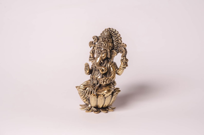 May include: A detailed brass statue of the Hindu deity Ganesha, seated on a lotus flower. The statue features intricate carvings, a large head with elephant features, and multiple arms. The figure is in a seated position with hands in a blessing gesture.