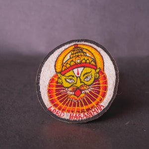 May include: Embroidered patch featuring a deity with a yellow face, orange and red headdress, and a red beard. The text "LORD NARASIMHA" is embroidered in red around the bottom edge of the patch. The patch is circular with a white background and black border.