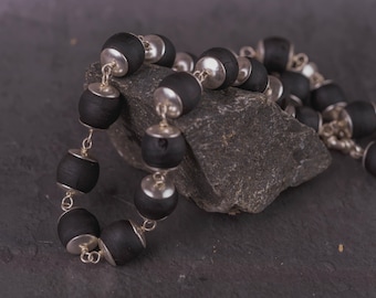 May include: A black beaded necklace with silver-coloured metal accents. The necklace features barrel-shaped black beads and small silver-coloured metal caps and connectors. The jewellery is displayed on a dark grey stone.
