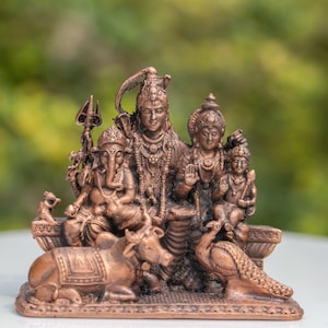 May include: A bronze-coloured sculpture depicting Hindu deities, including Shiva, Ganesha, and others. The detailed figures are arranged on a base, with a cow and peacock. The background is a blurred green.